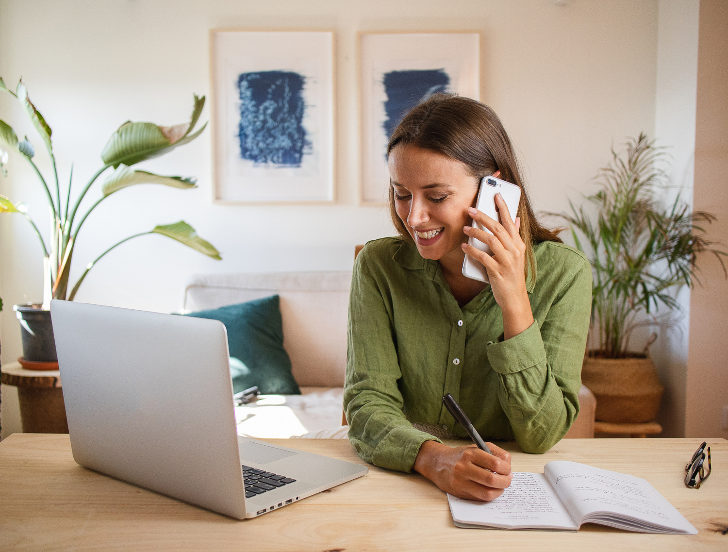 Femme souriante au téléphone prenant des notes devant son ordinateur, illustrant l'accompagnement personnalisé pour les aidants et les personnes en perte d'autonomie.